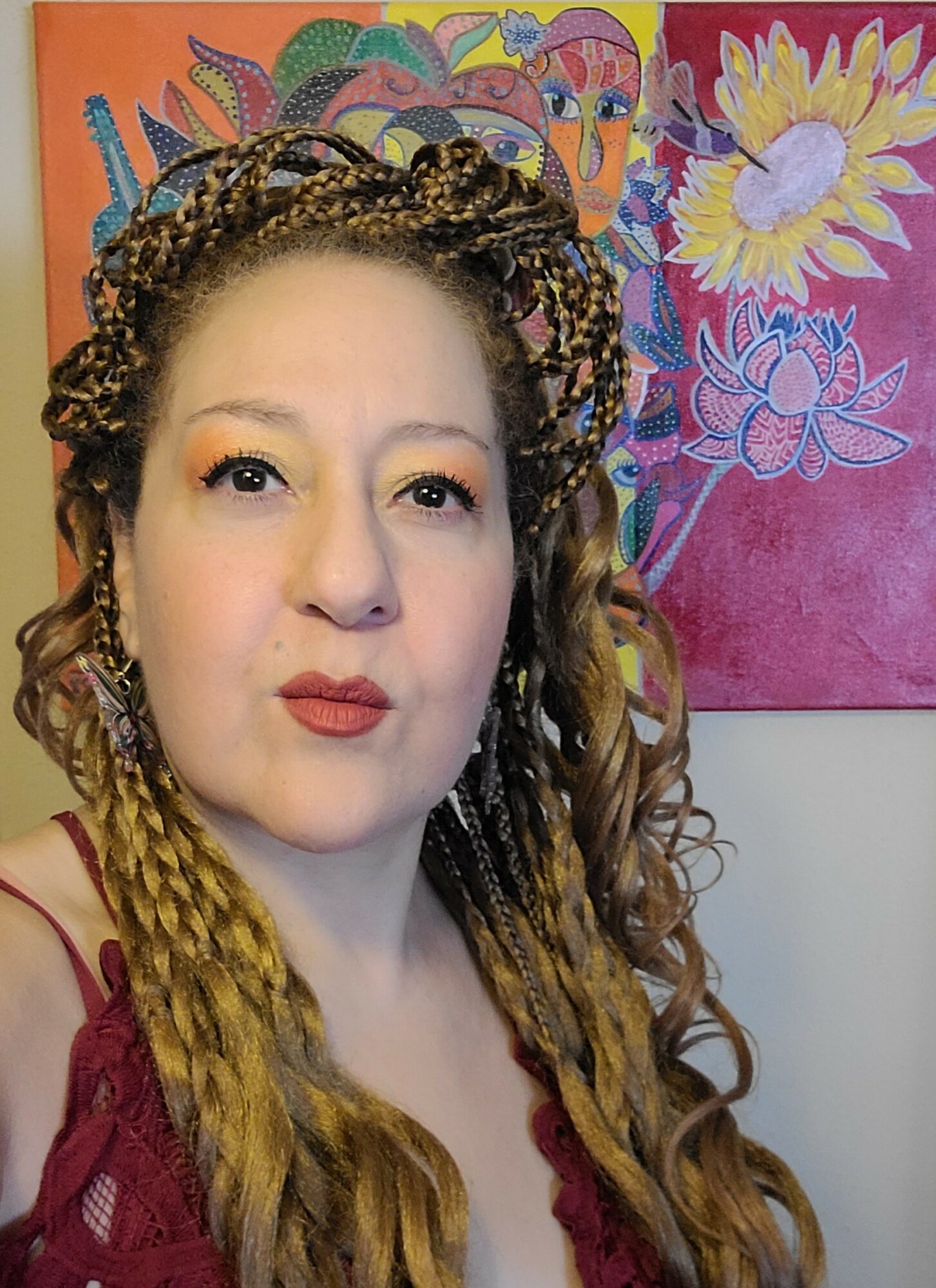 Woman with braided hair and makeup, wearing a red top, standing in front of colorful floral artwork.