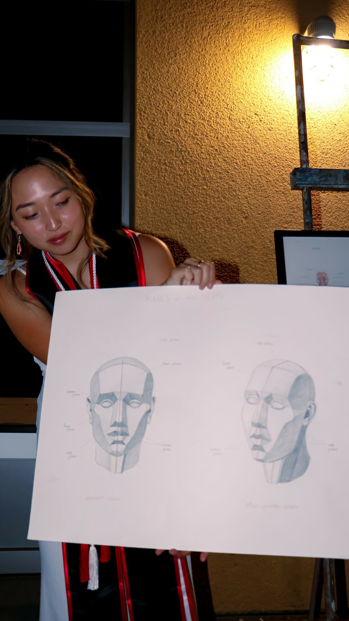 Young woman holding a drawing of two faces, standing outside at night near a yellow wall and a lit lamp.