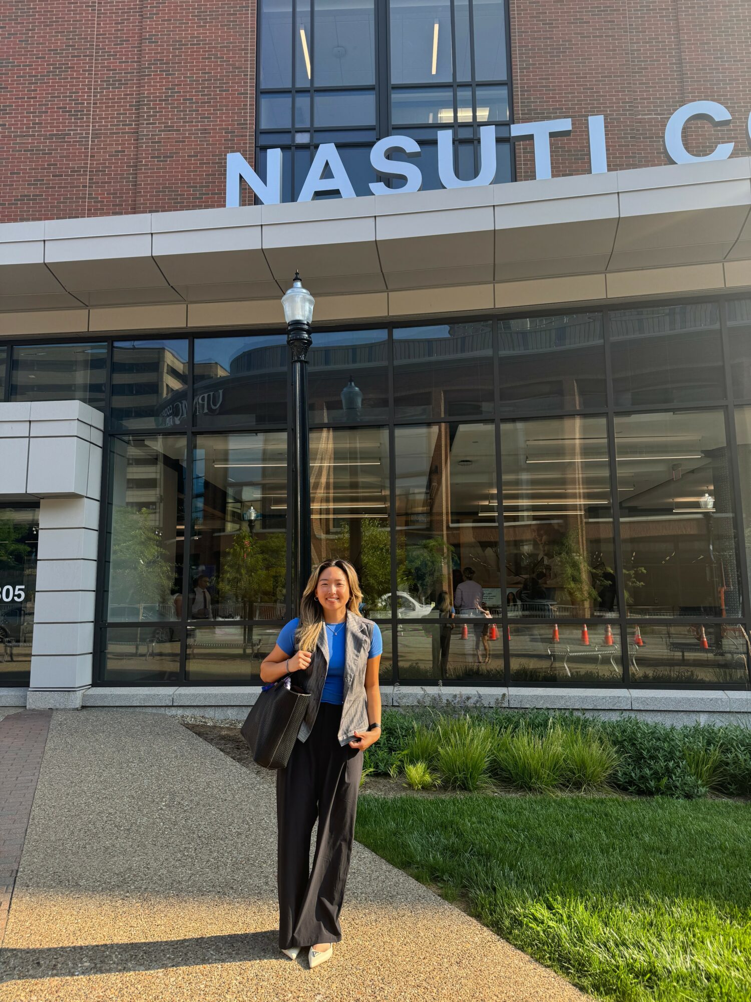 Woman standing outside a modern building with large glass windows and a sign reading NASUTI C, holding a bag and smiling.