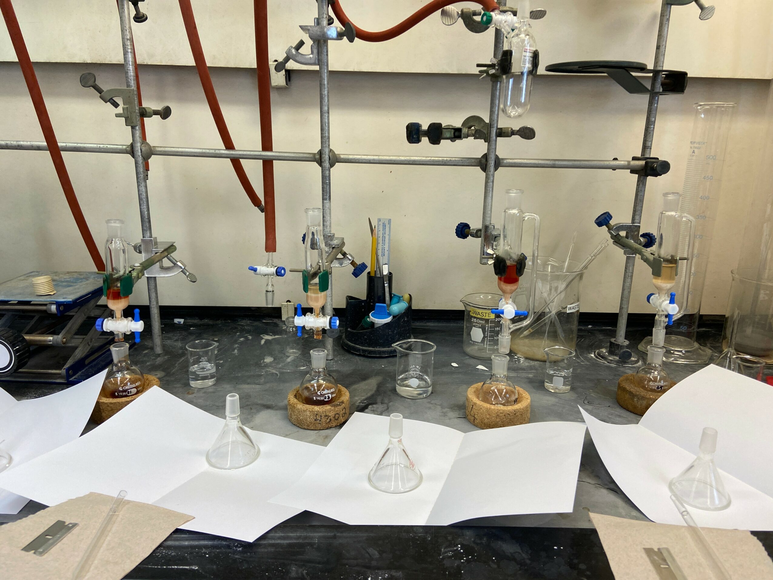 Laboratory setup with glassware, metal stands, and rubber tubing on a work surface, with papers and containers nearby.