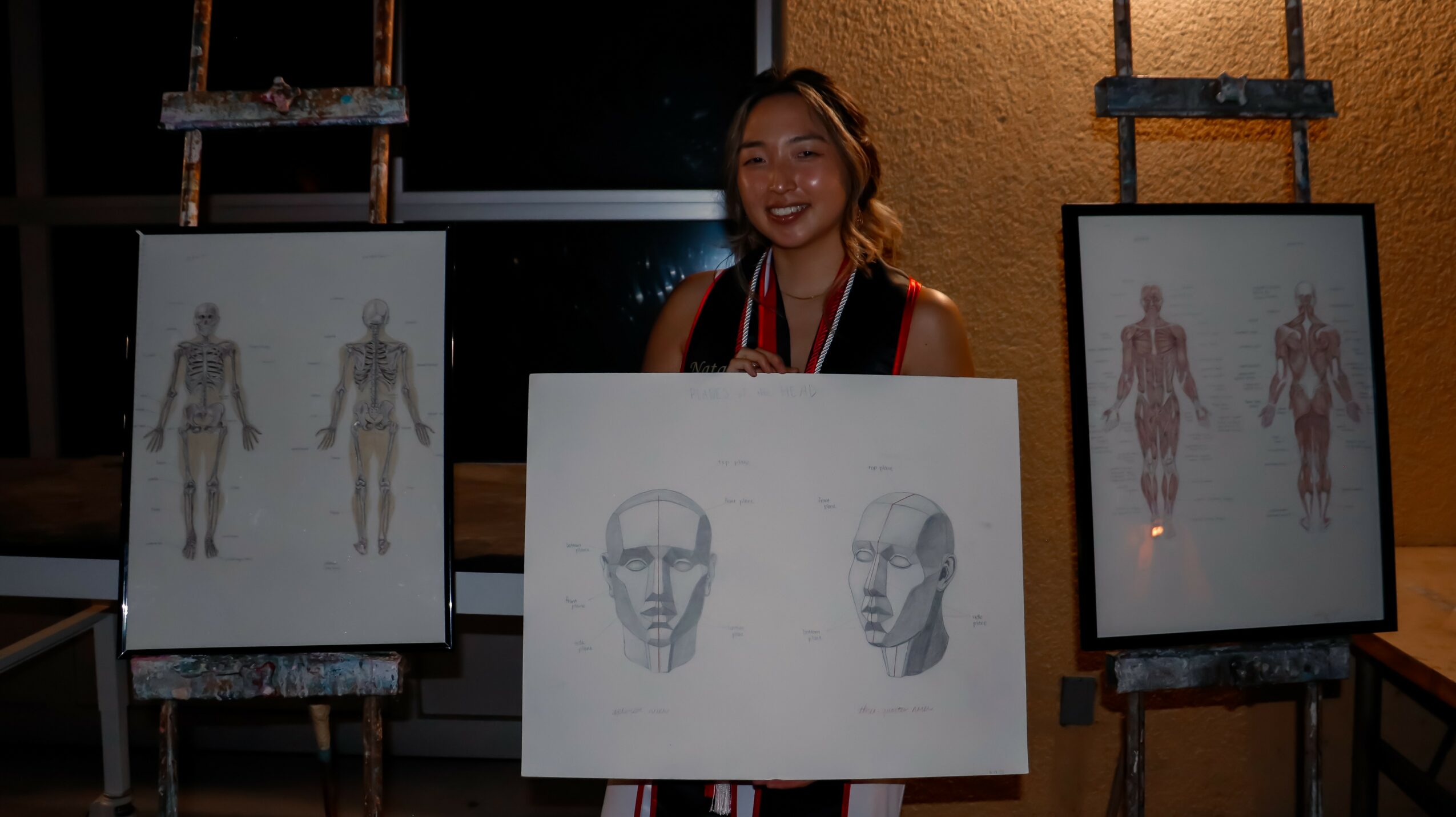 Young woman standing behind a drawing of a face, with anatomical posters on easels on either side, in a dimly lit room.