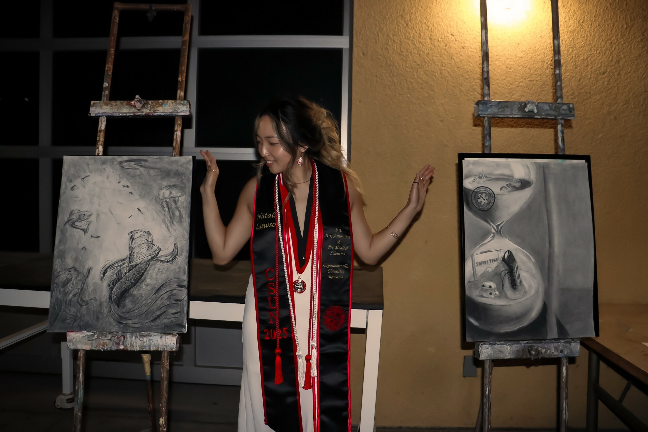 Person standing between two paintings on easels, wearing a black outfit and a red and black scarf, smiling.