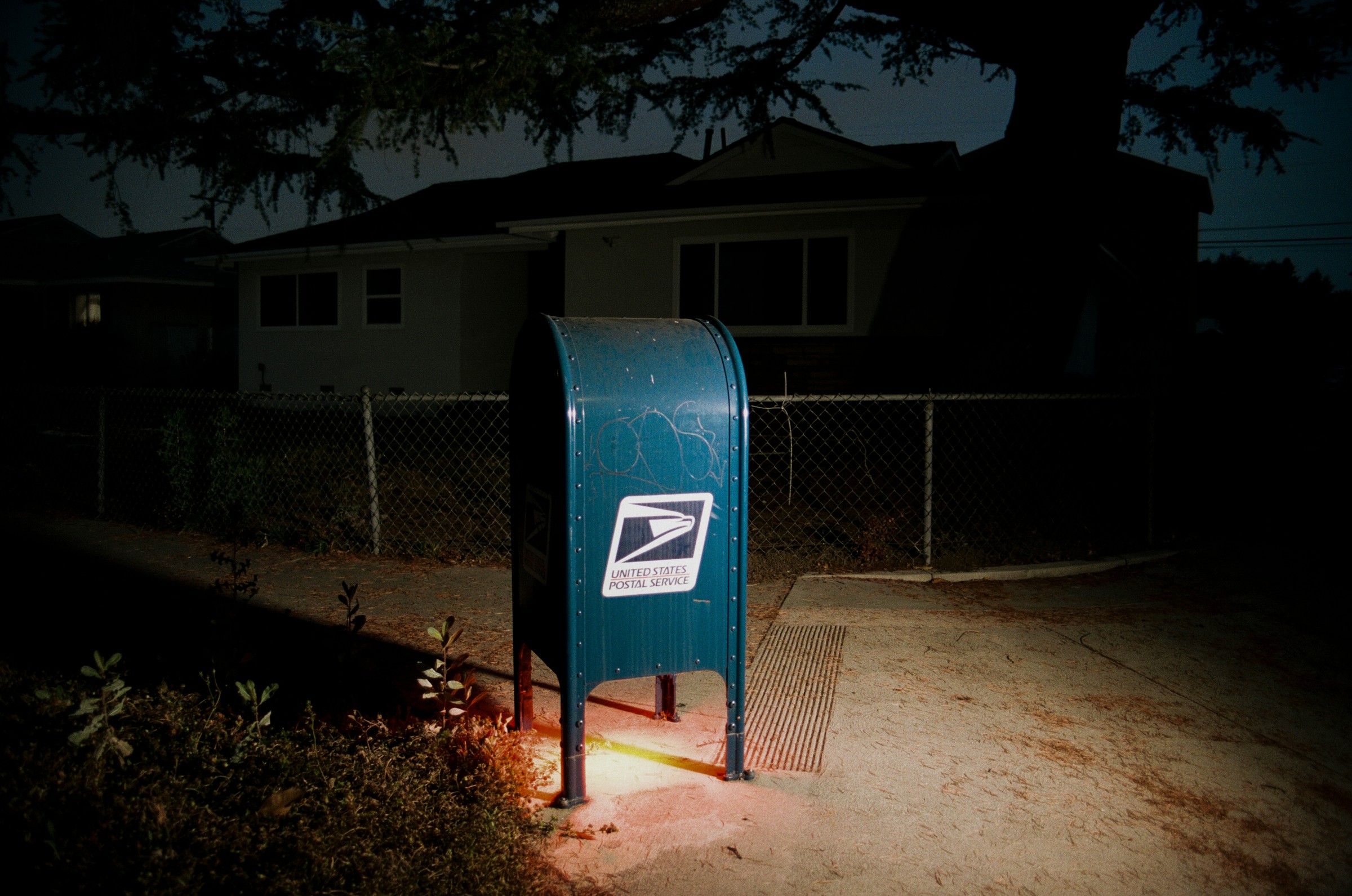 Blue mailbox with USPS logo on a sidewalk at night, illuminated by nearby light, with a house and fence in background.