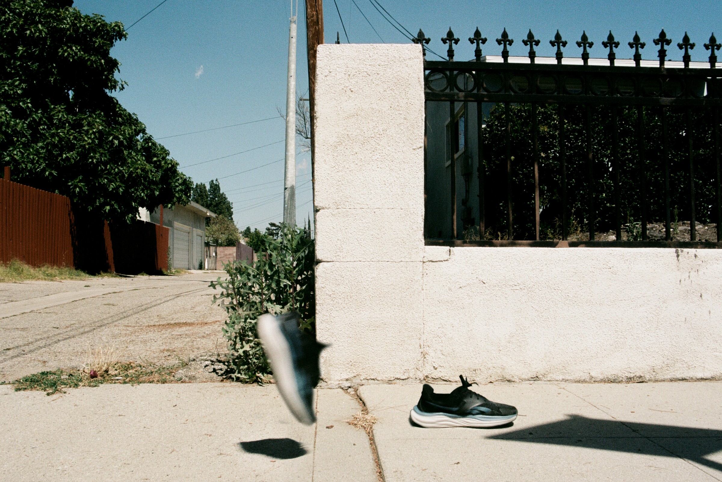 Pair of shoes on sidewalk near a white pillar and black fence, with a street and trees in background.