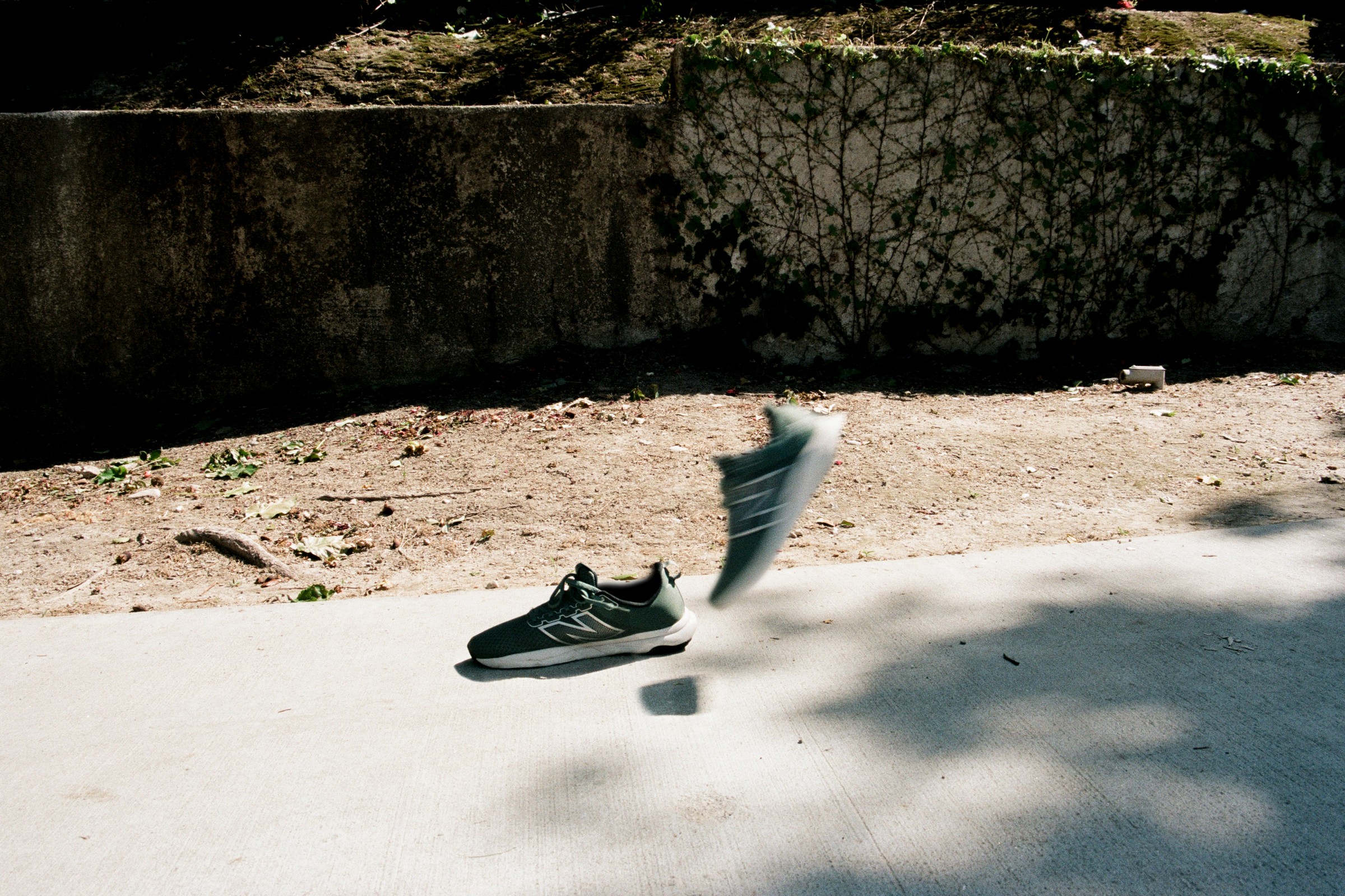 A single sneaker and a sock are on the ground outdoors, with shadows and a wall in the background.