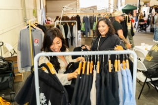 Two women browsing clothing racks at an outdoor market, smiling and shopping for pants, with other vendors and shoppers in background.