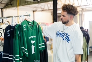 Man with curly hair wearing a white shirt shopping for green and black jerseys in a store.