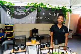Smiling woman standing behind a table with skincare products at a booth, banner reads 'Branch + Vine Skincare Co.'