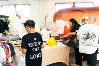 People shopping at a market stall with a large orange food truck in the background, some browsing clothes and others buying items.