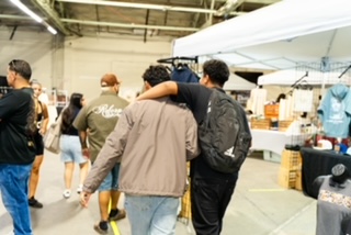 Two people walk together, one with arm around the other's shoulder, in an indoor market or event space.