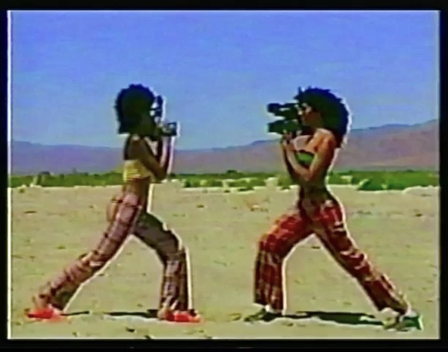 Two children in plaid pants and tank tops face each other in a desert landscape, each holding a video camera, filming each other.