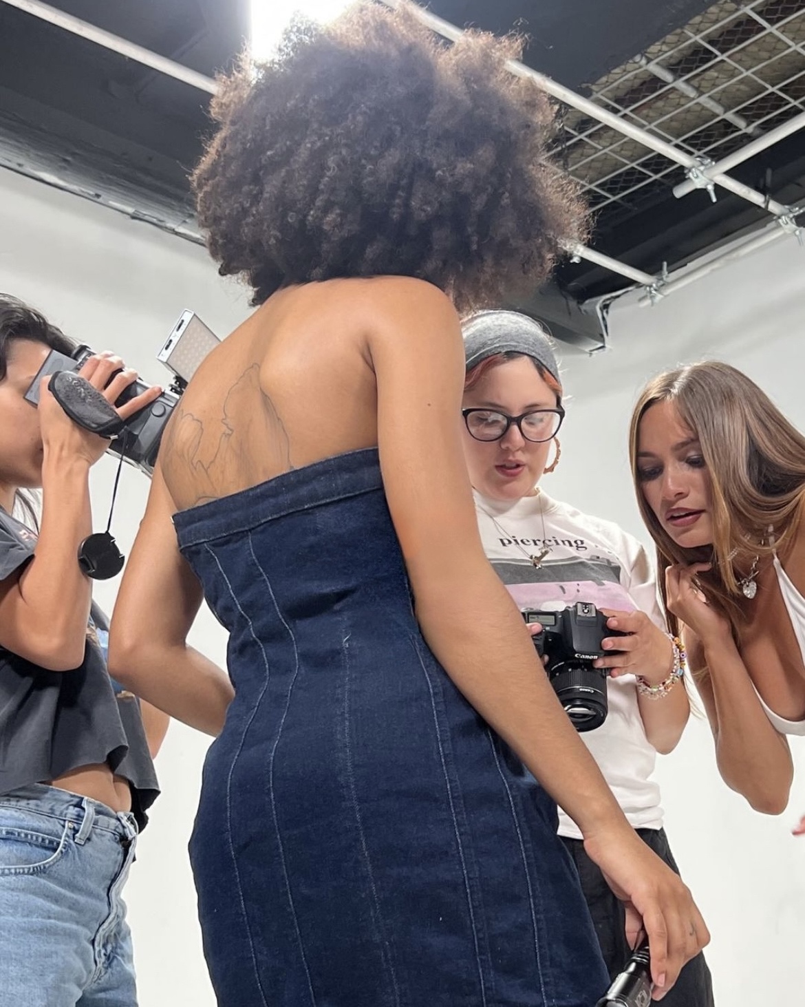 Three women in a studio, one with curly hair in a strapless dress, two with cameras, discussing something.