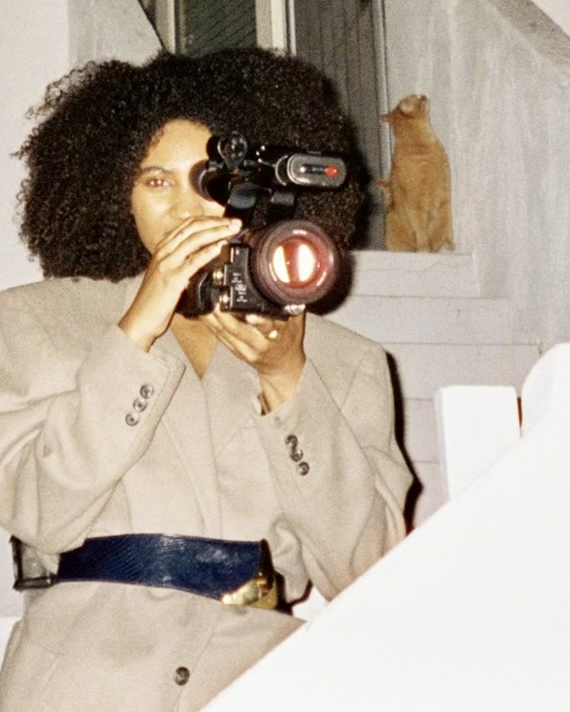 Person with curly hair holding a camera, wearing a beige blazer with a dark belt, in front of a window with a cat on the sill.