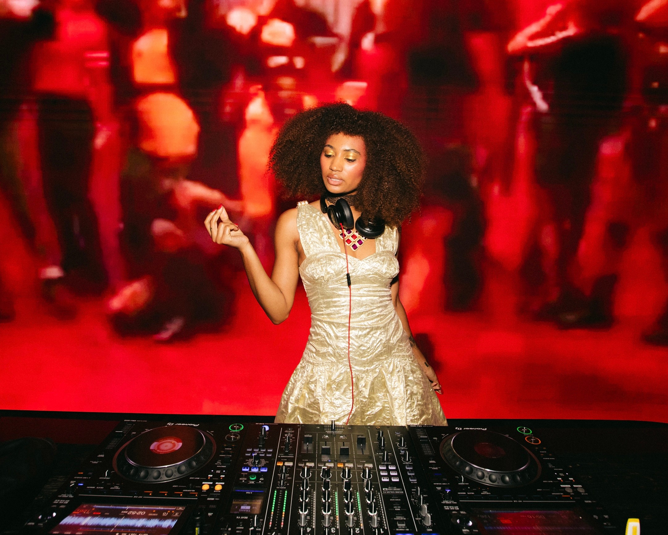 A woman with curly hair wearing a gold dress and headphones, DJing at a party with a red background.