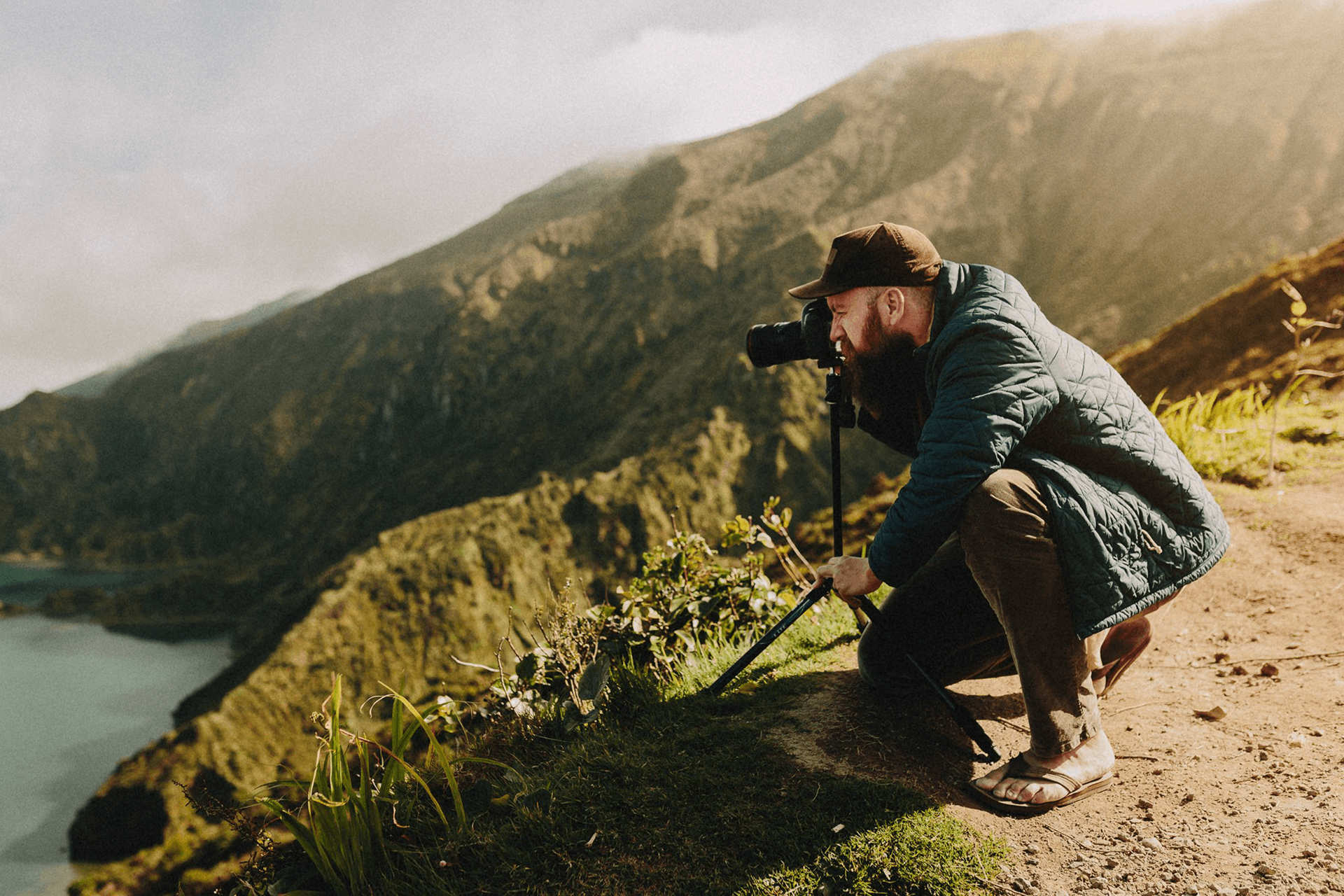 Person crouching on a trail with a camera on a tripod, overlooking a mountain landscape with a body of water, sunny weather