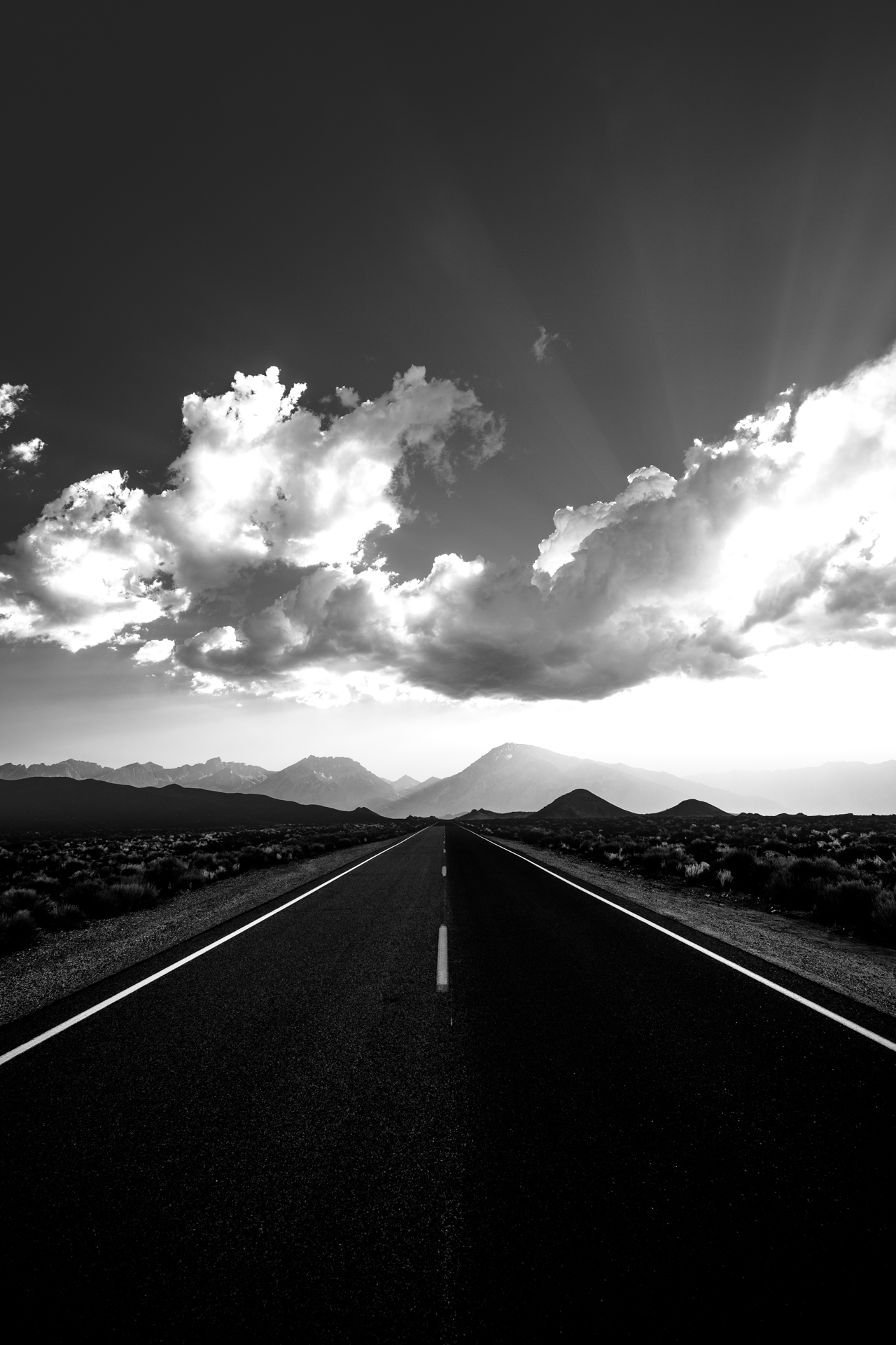 Long empty road stretching toward mountains under cloudy sky with sunlight rays, black and white image.