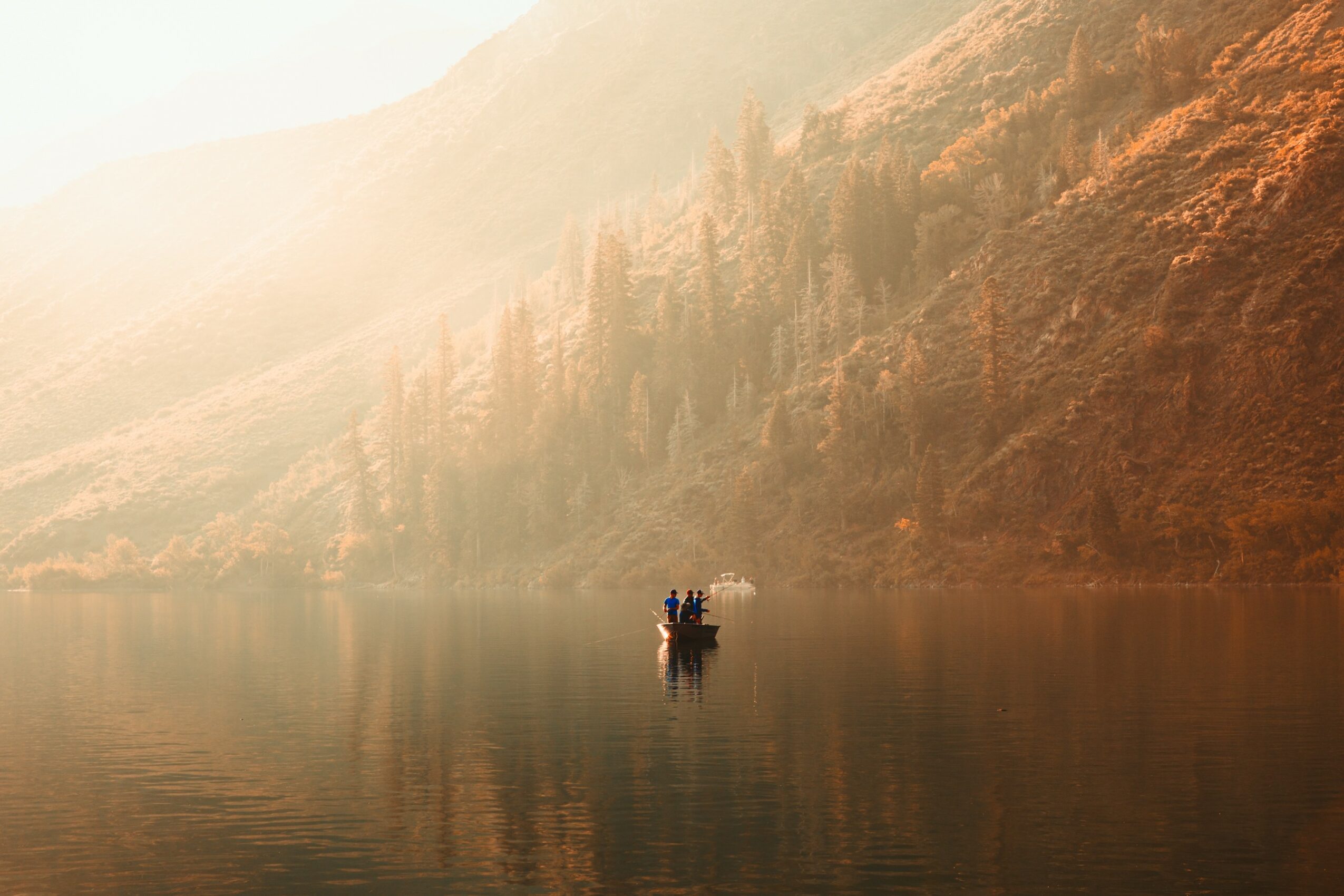 A boat with people on a calm lake surrounded by mountains and mist, with reflections on the water.