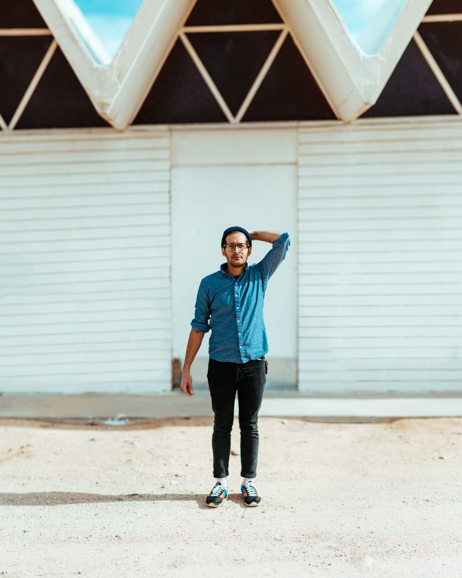 Young man standing outdoors in front of a white wall, wearing glasses, a blue jacket, black pants, and sneakers, with one hand on his head.