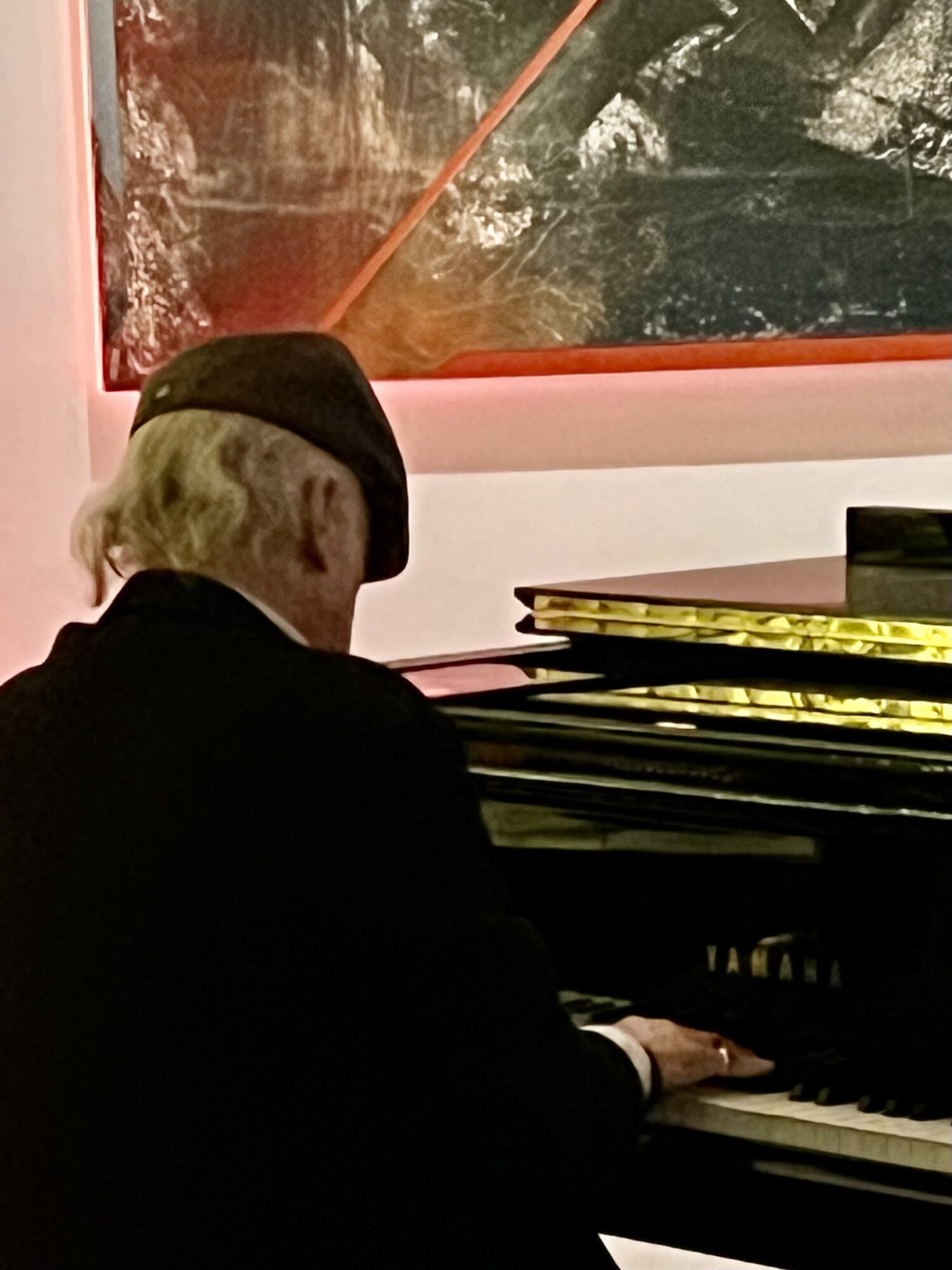 Person with light hair wearing a black hat playing a Yamaha piano, with a colorful abstract painting in the background.