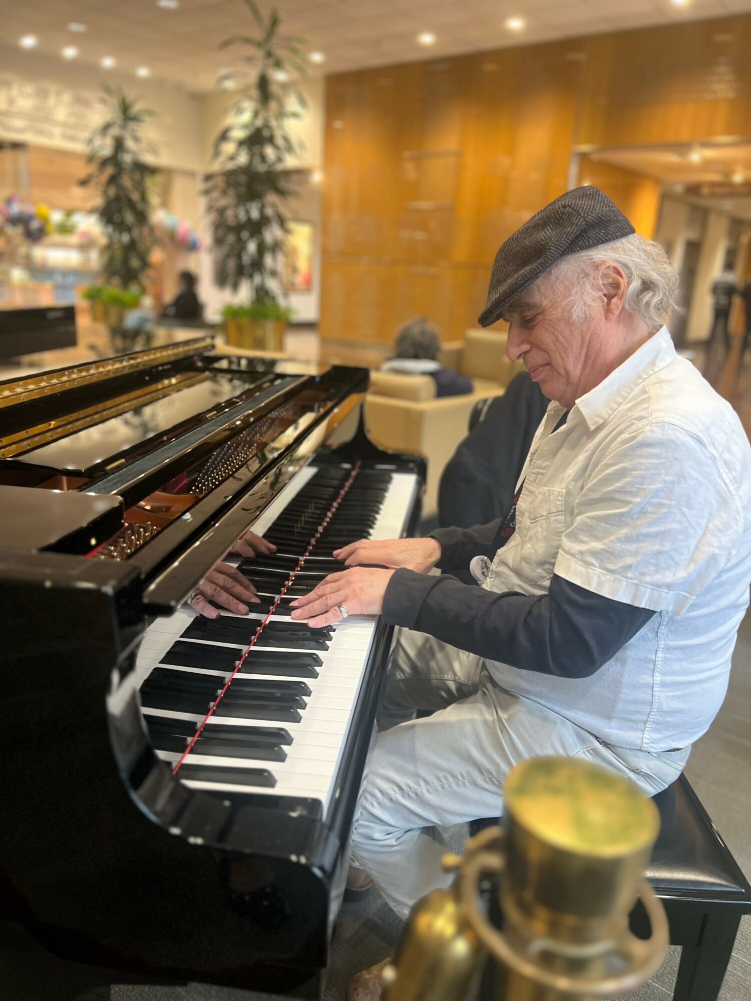 Older man playing piano in a hotel lobby, wearing a white shirt, black cap, and black sleeves, with plants and warm lighting.