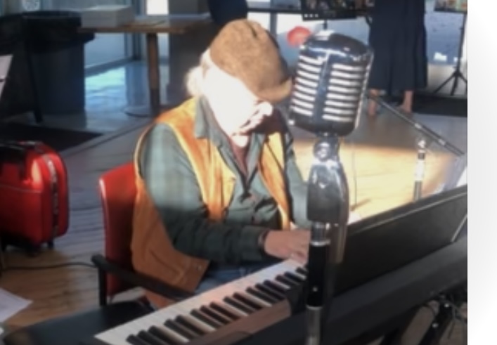 Person playing piano at a table with a microphone, wearing a hat and orange vest, in a room with large windows.