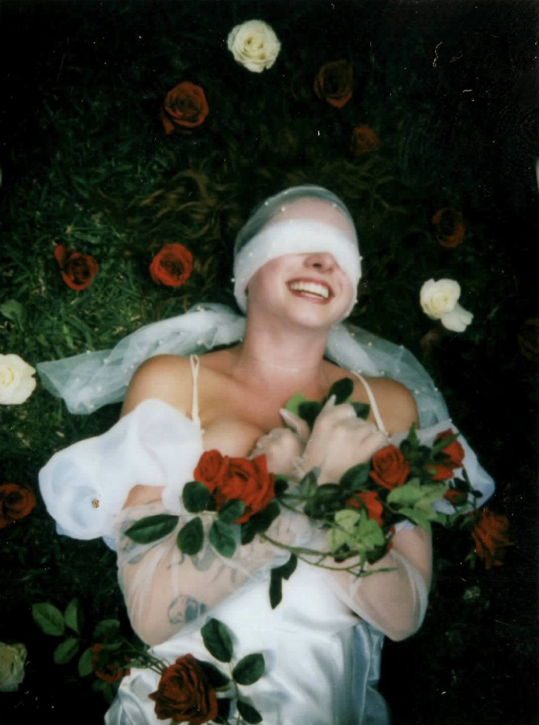 Woman lying on grass surrounded by red and white roses, smiling with eyes covered by a blindfold, holding a rose.