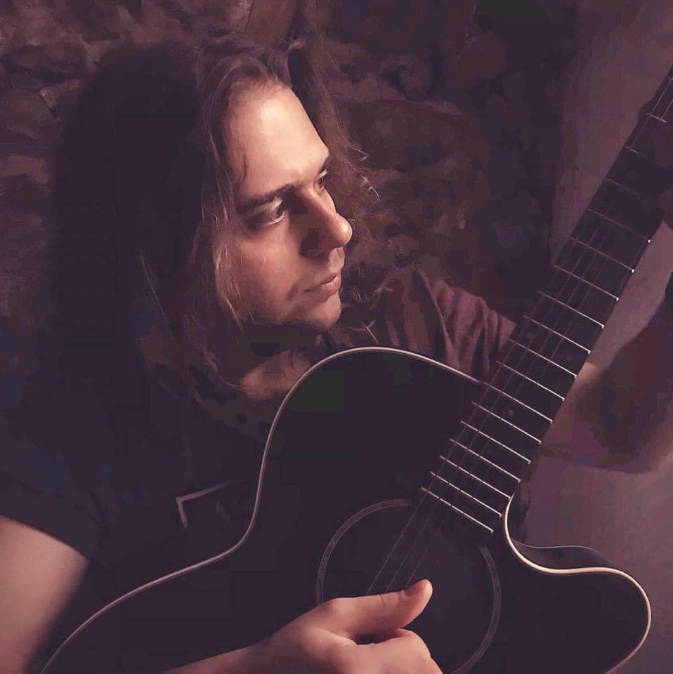 Person with long hair holding a black acoustic guitar, looking to the side in a dimly lit setting.