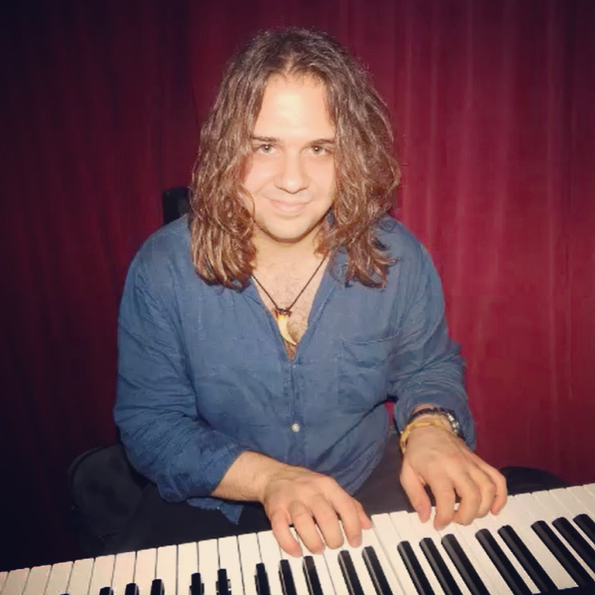 Person with long wavy hair playing a keyboard, wearing a blue shirt, in front of a red curtain background.
