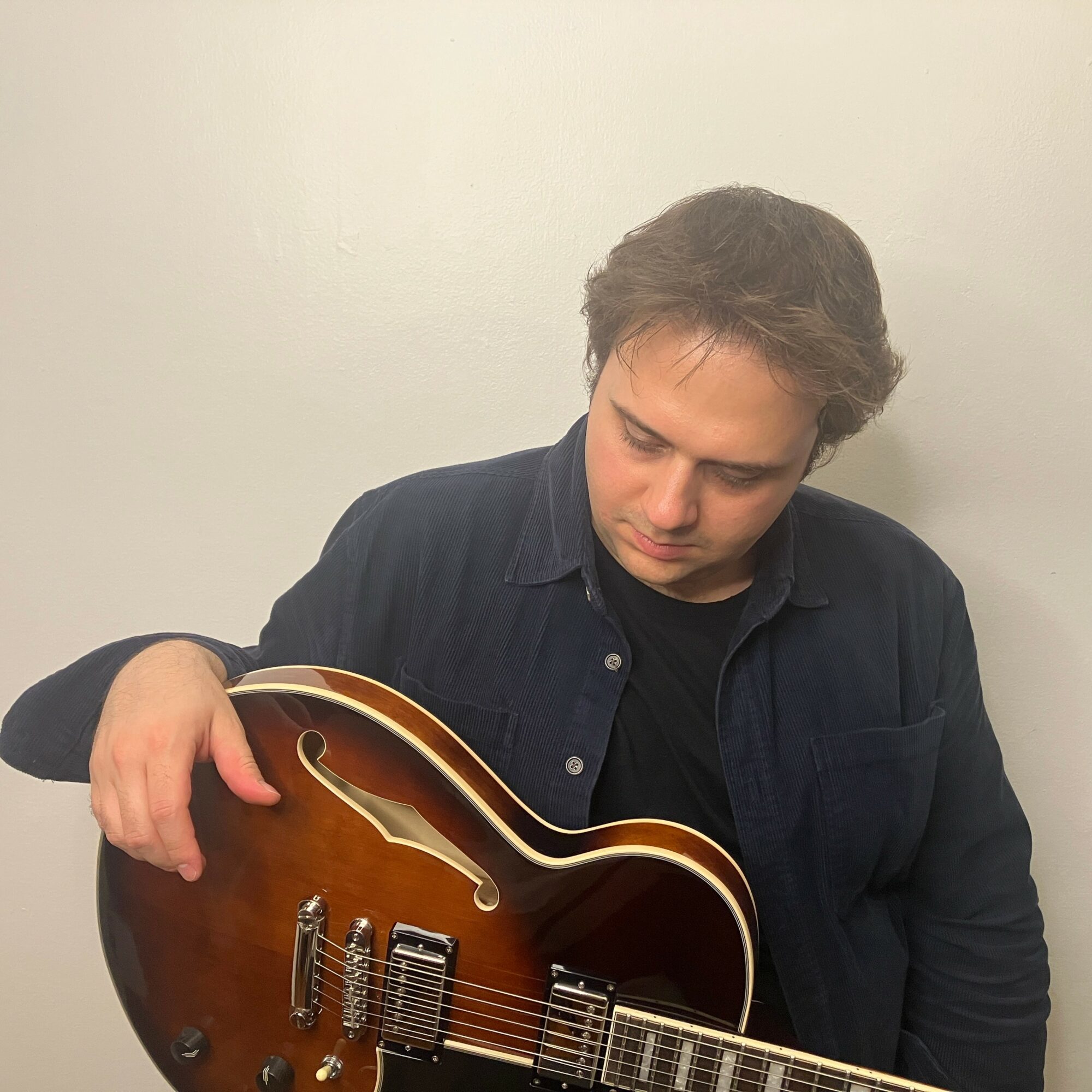 Young man with dark hair wearing a dark shirt, holding a guitar, looking down at it, against a plain light background.