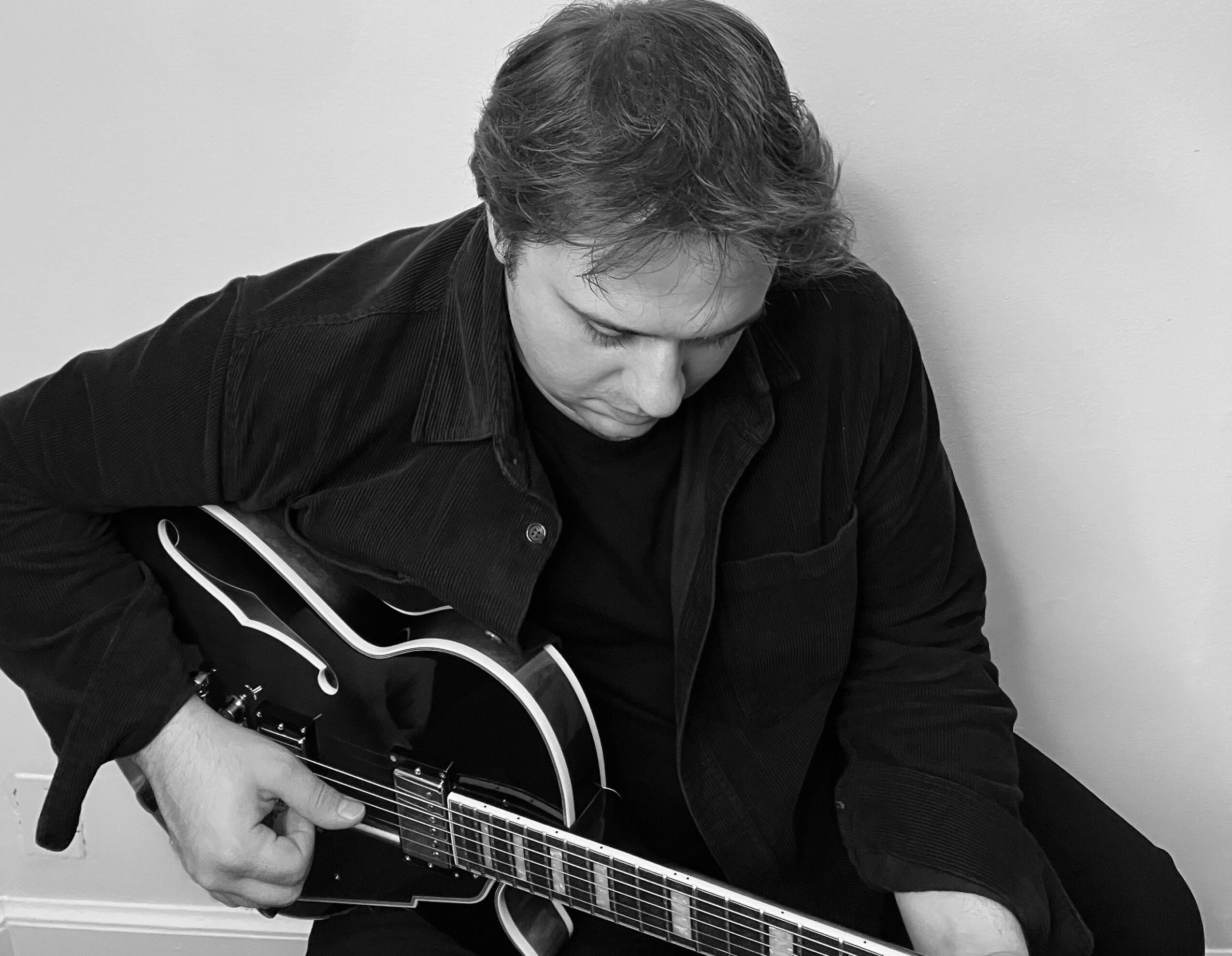 Person with medium-length hair playing an electric guitar, looking down, wearing a dark shirt, against a plain background.