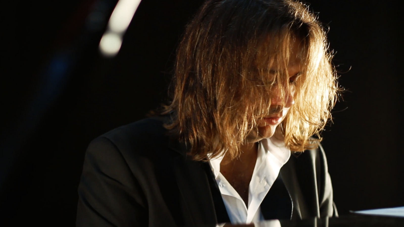 Person with shoulder-length hair looking downward, wearing a dark blazer and white shirt, in a dark setting.