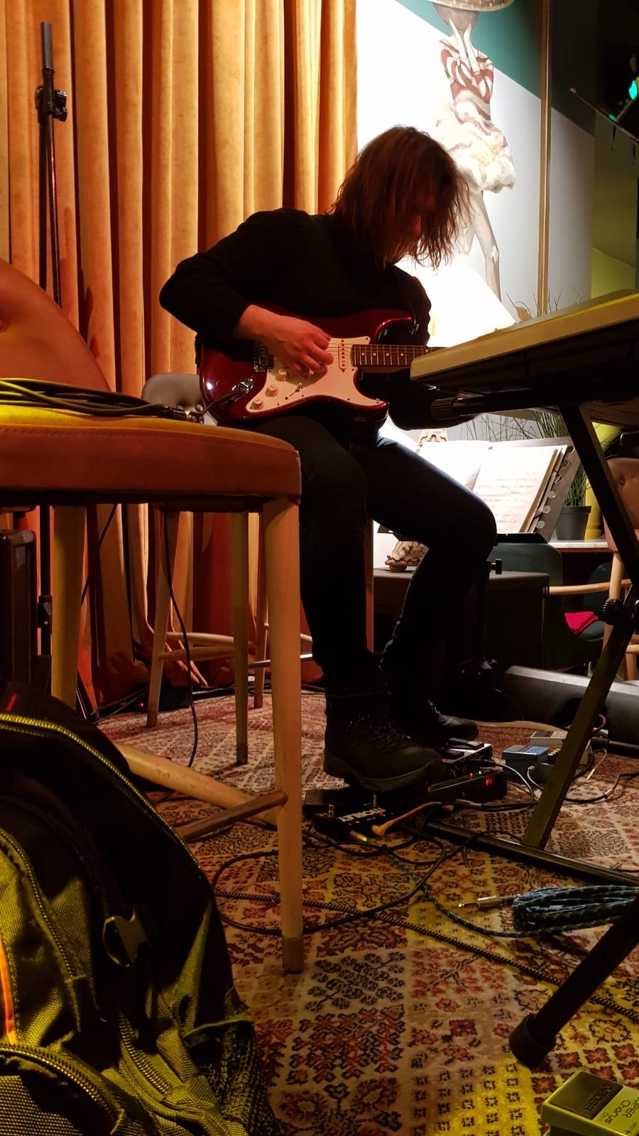 Person with curly hair playing electric guitar in a room with curtains and musical equipment.