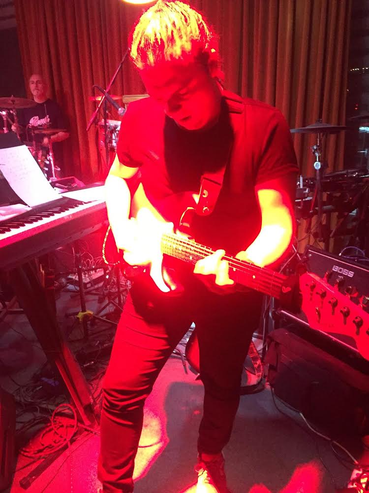 Person playing guitar on stage with red lighting, surrounded by musical equipment and a keyboard, curtain backdrop.