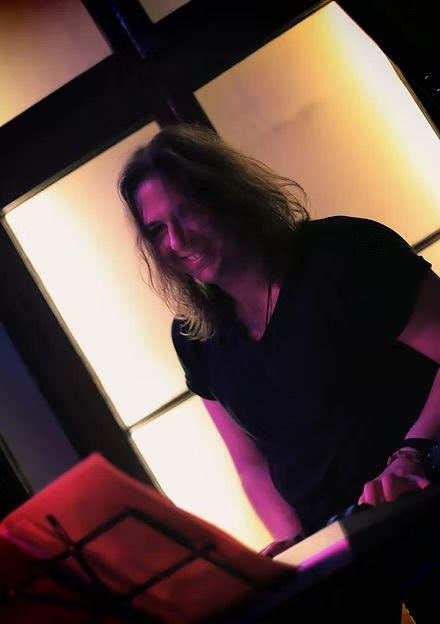 Person with shoulder-length hair smiling, sitting in front of illuminated windows, wearing a black shirt, with a music stand nearby.