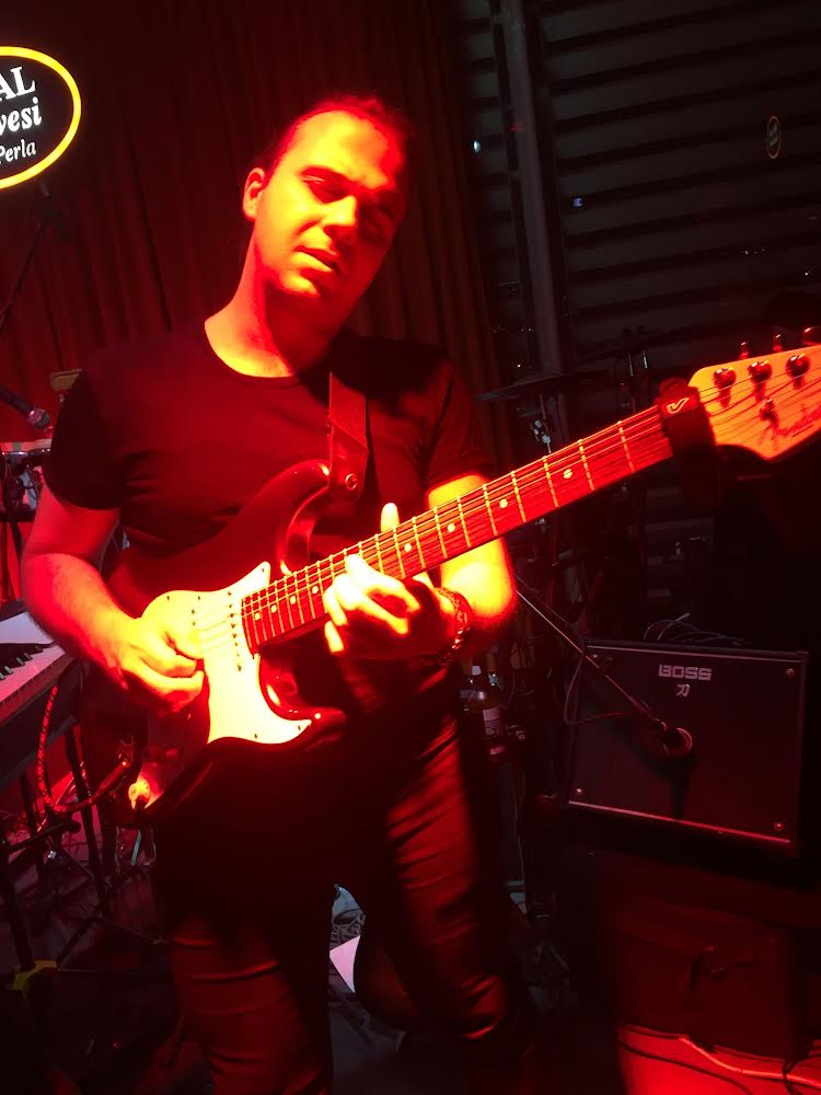 Person playing electric guitar on stage with dark background and equipment, illuminated by red lighting.