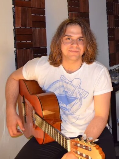 Person with shoulder-length hair playing an acoustic guitar, wearing a white t-shirt, sitting indoors.