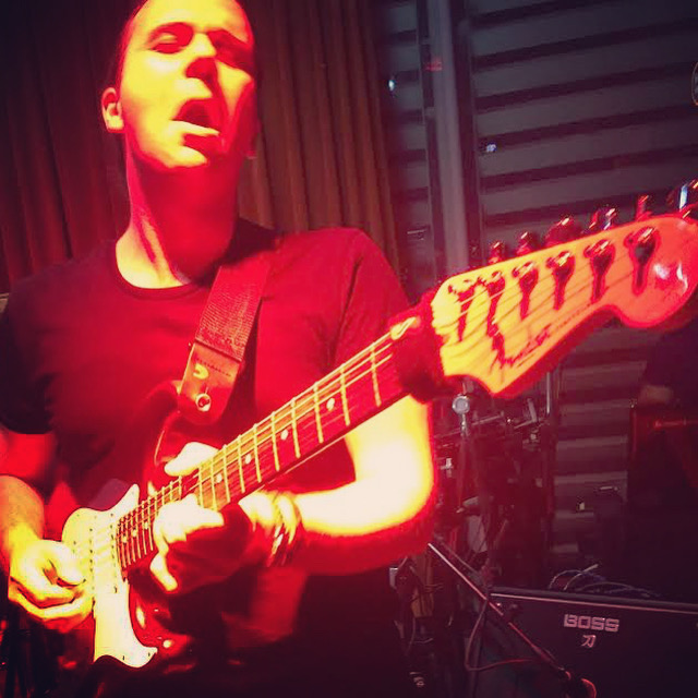 Person playing electric guitar with eyes closed, red lighting, dark background, and musical equipment visible.
