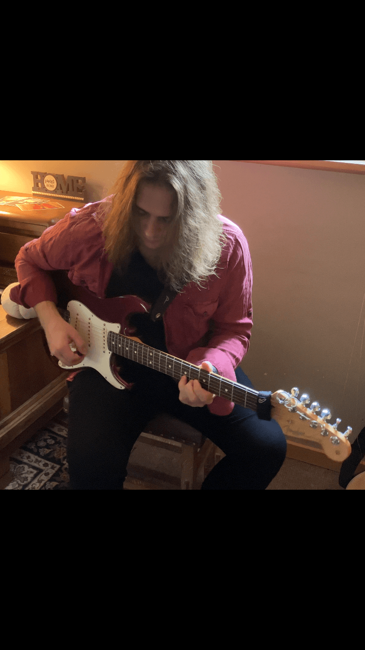 Person with curly hair playing an electric guitar indoors, sitting on a chair, with a wooden cabinet in the background.