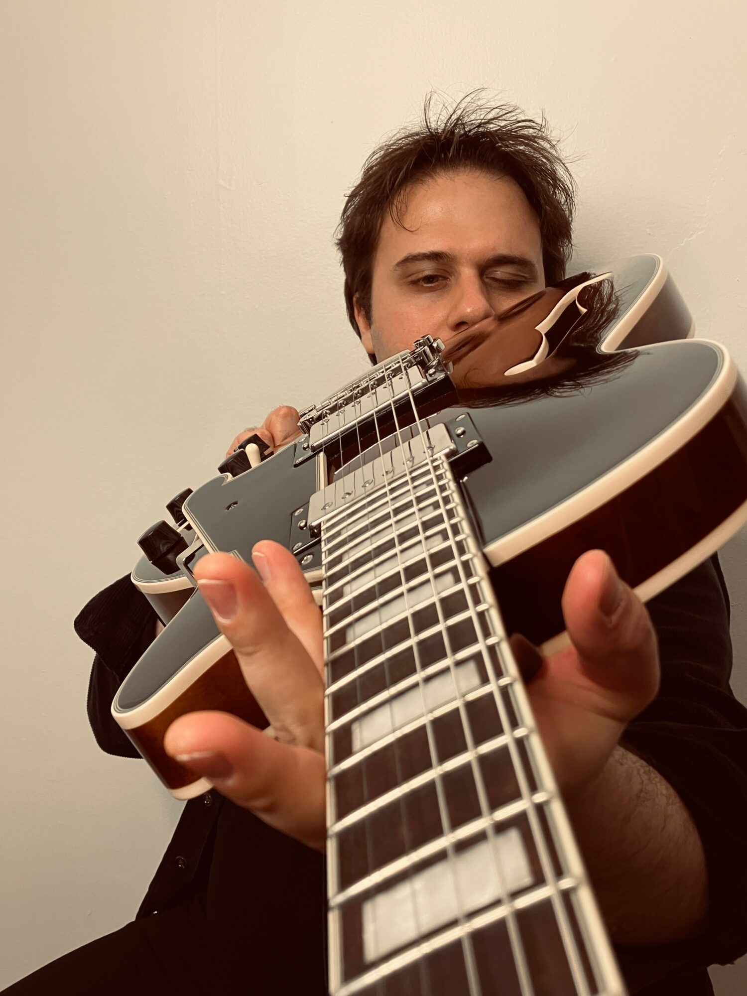 Person playing an acoustic guitar, looking down at the fretboard, with a plain light-colored wall background.