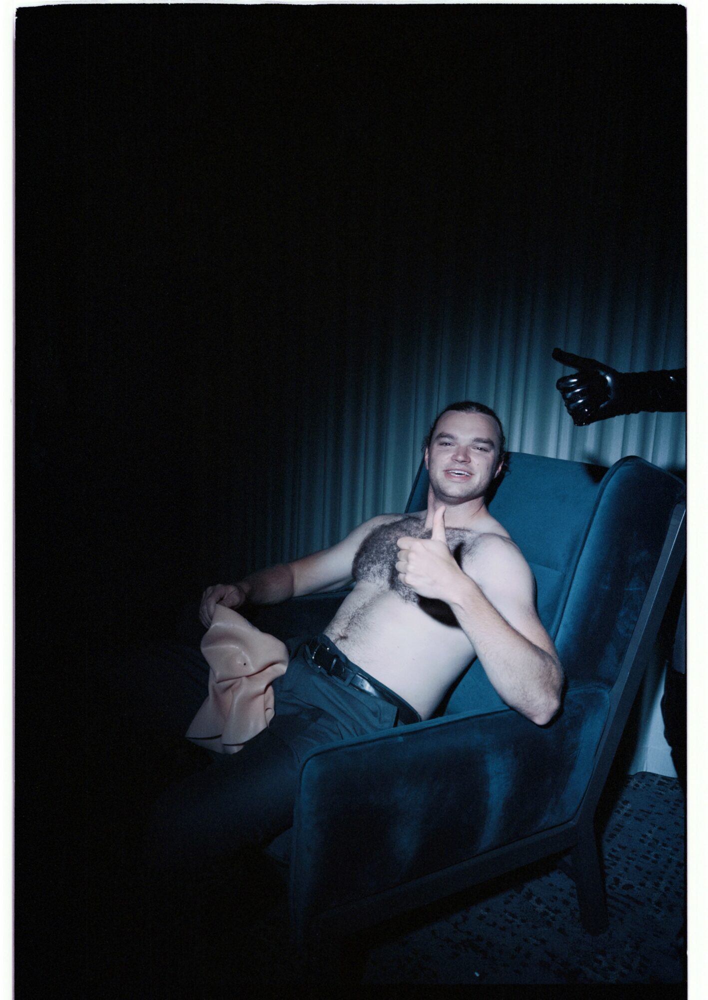 Young man reclining shirtless in a chair, with a robotic arm reaching towards him, dark background, blue lighting