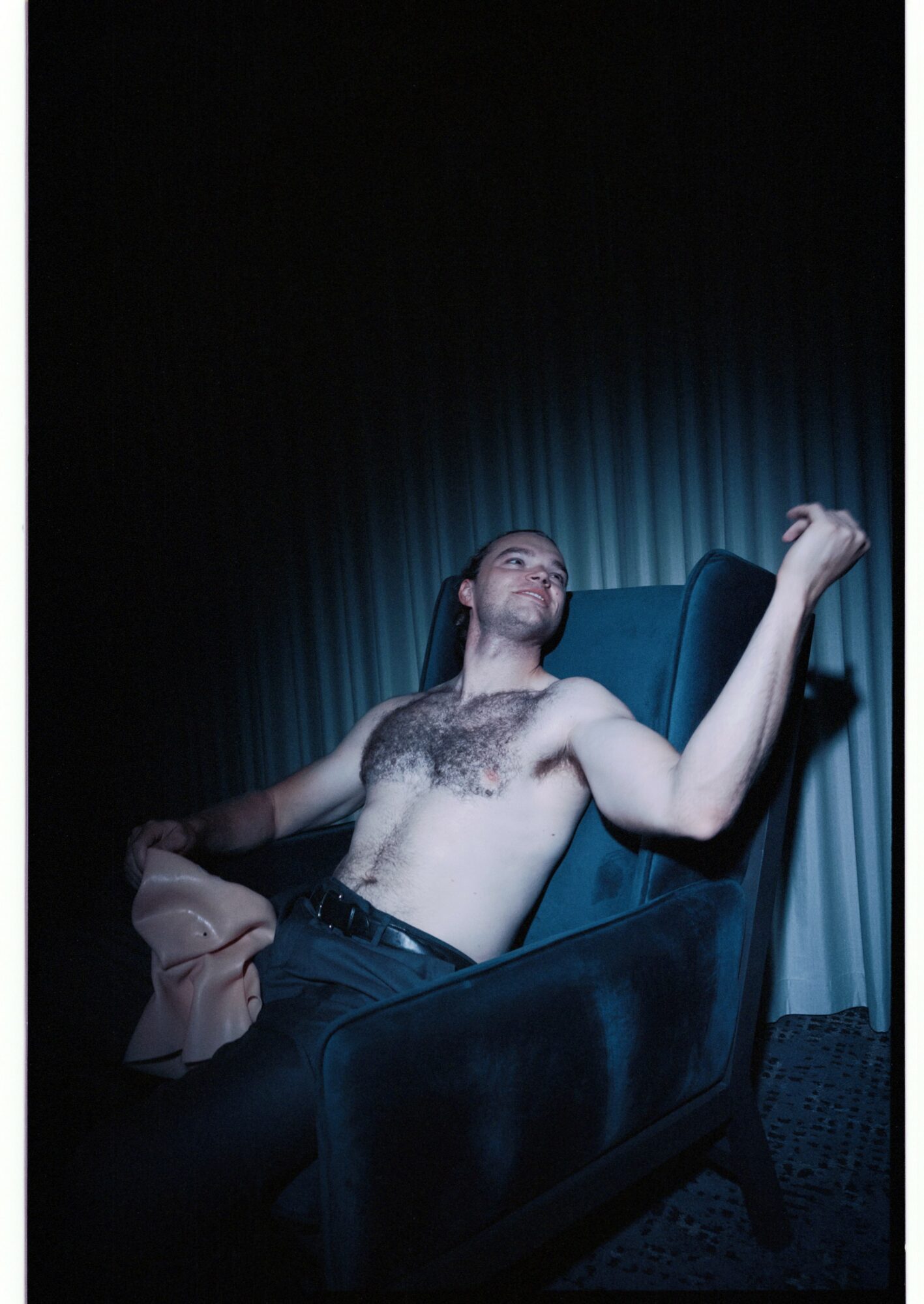 Man reclining shirtless on theater seat with curtain backdrop, holding a cloth, looking upward.