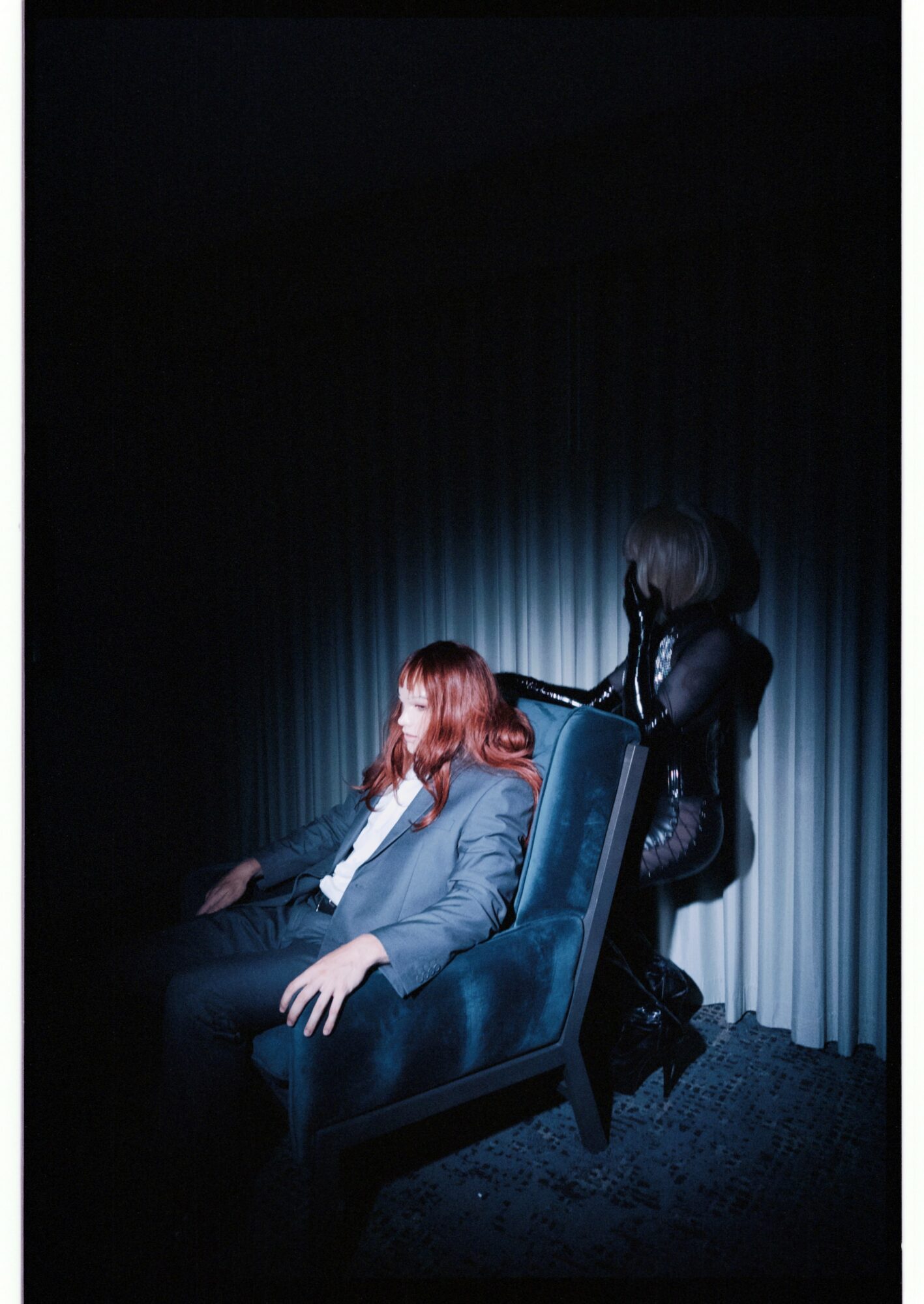 Person with red hair sitting in a chair, another person standing behind, both in a dark room with curtains.