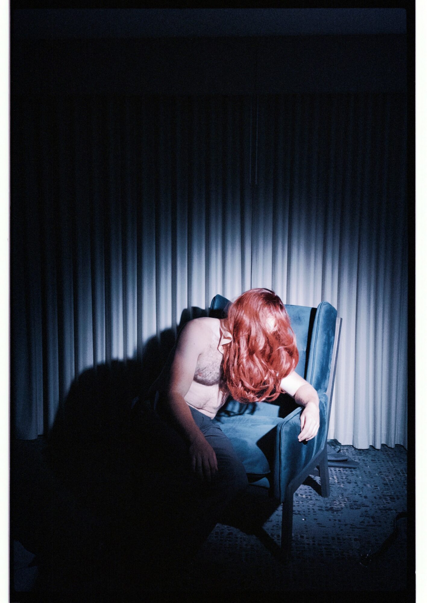 Person with red hair leaning over a chair in a dimly lit room with curtains, head down, face not visible.