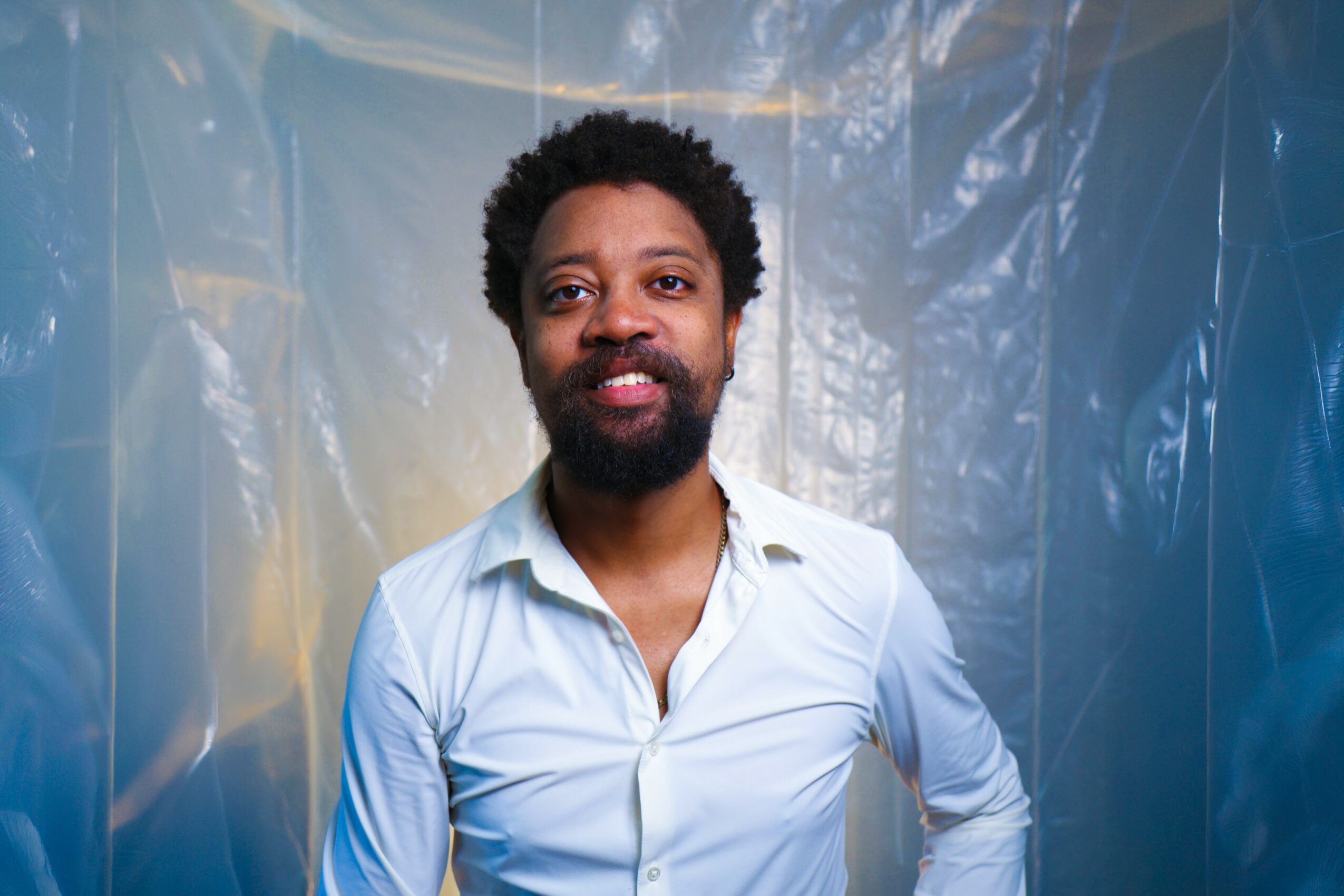 Man with curly hair and beard wearing a light blue shirt, smiling, standing in front of a blue plastic background.