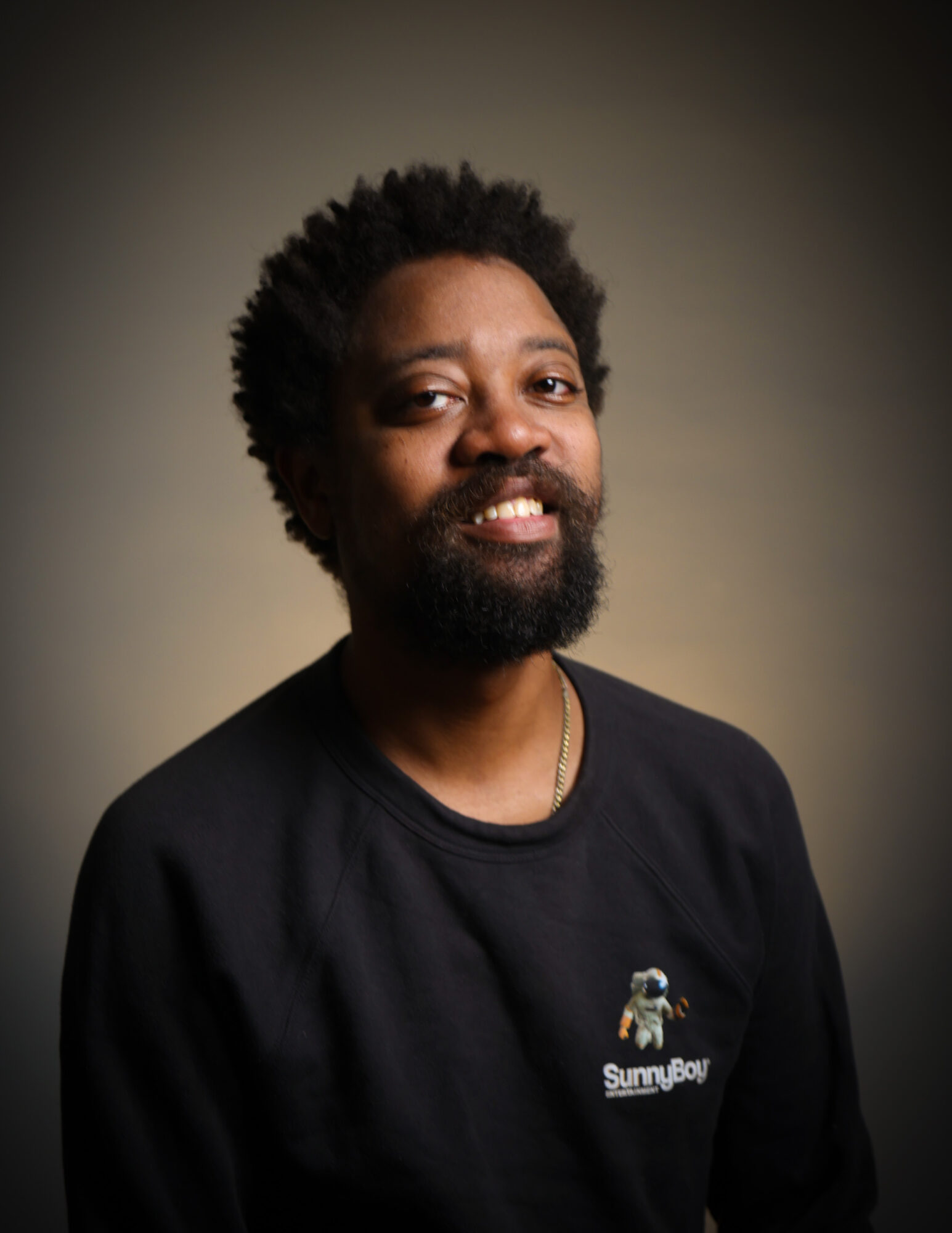 Portrait of a man with curly hair and a beard, wearing a black sweatshirt with a small logo, smiling slightly.