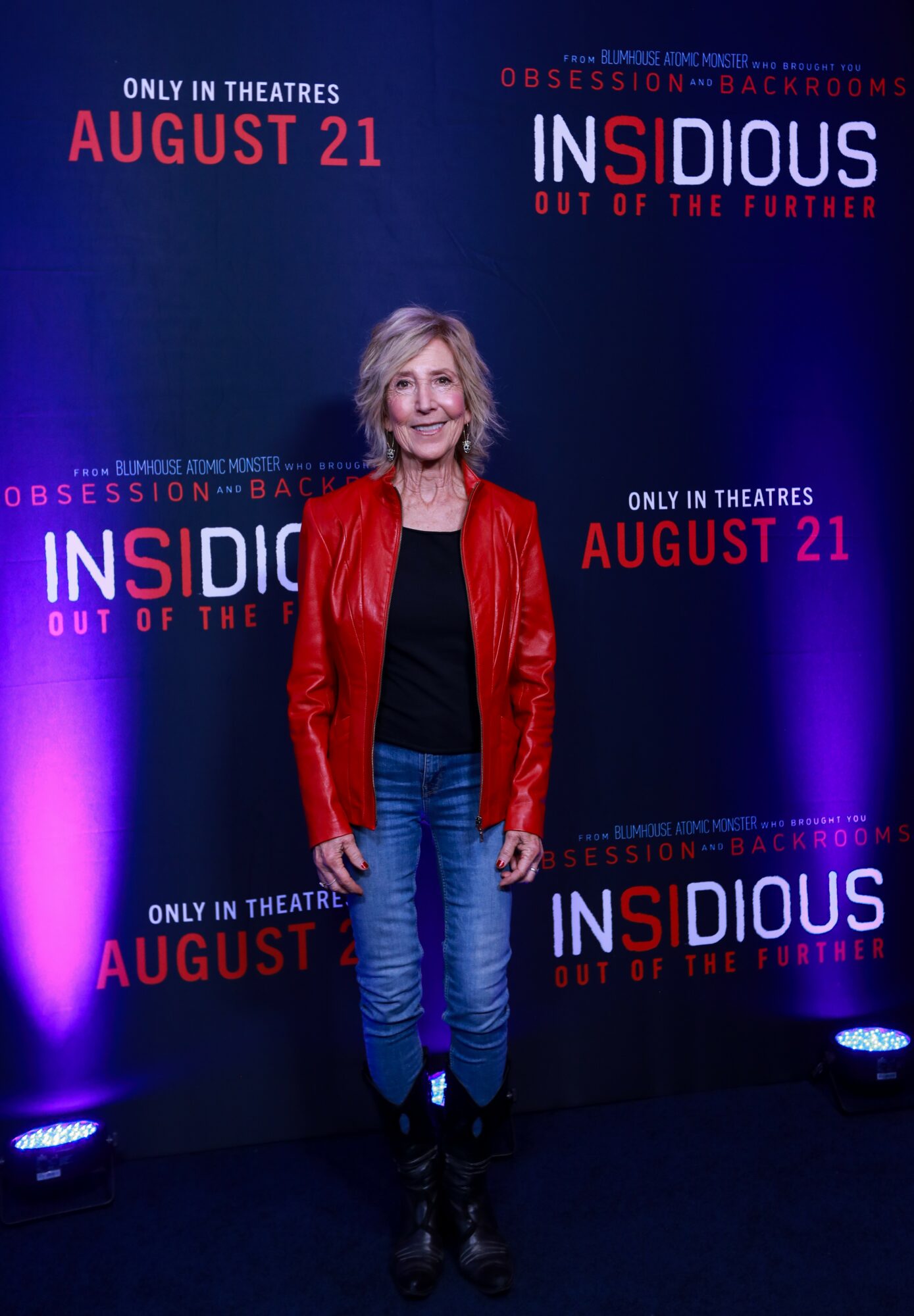 Woman with blonde hair in a red jacket, black top, and jeans standing in front of a dark promotional backdrop for 'Insidious: Out of the Forth' movie, August 21 release date.