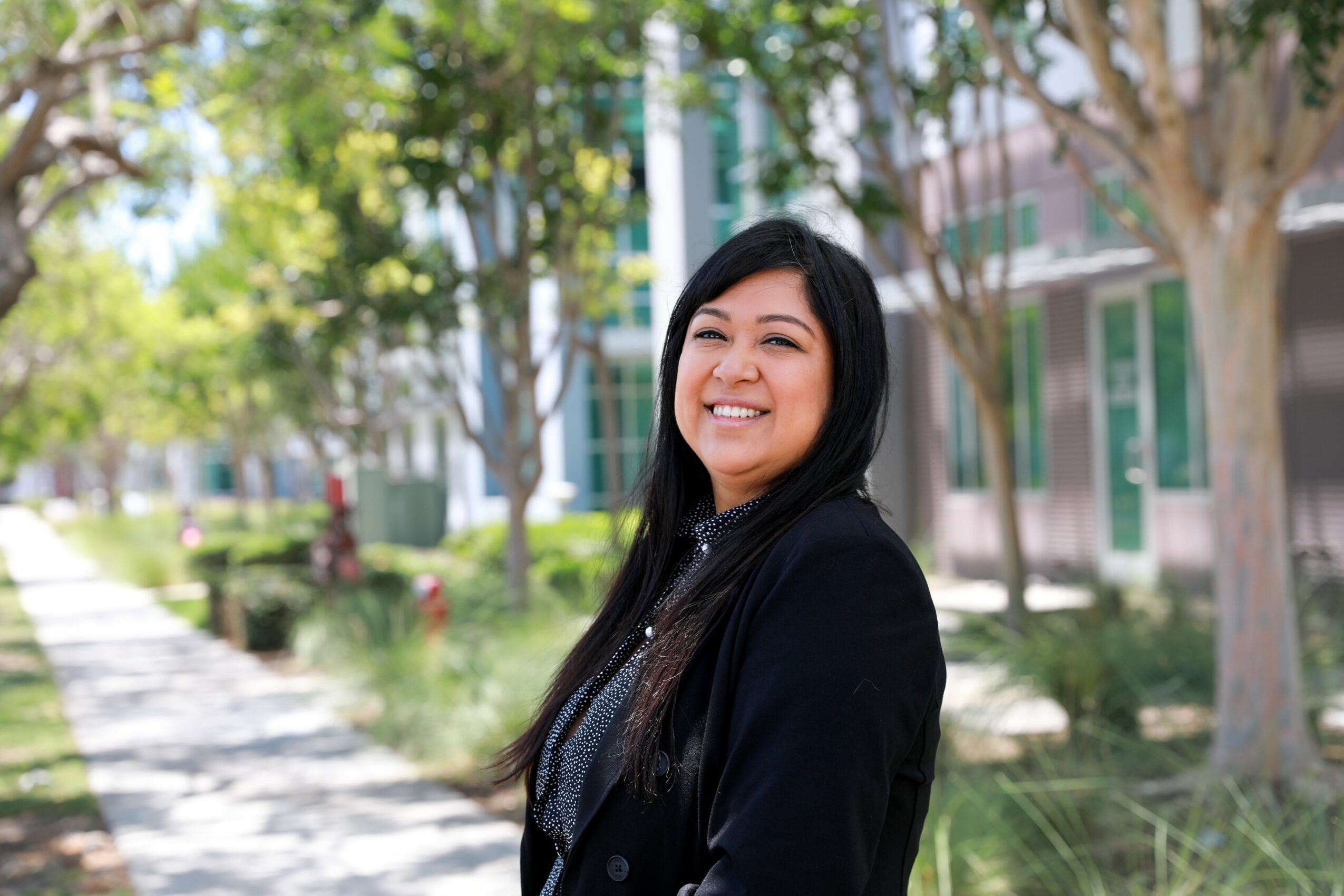 Smiling woman with long dark hair outdoors, trees and buildings in background