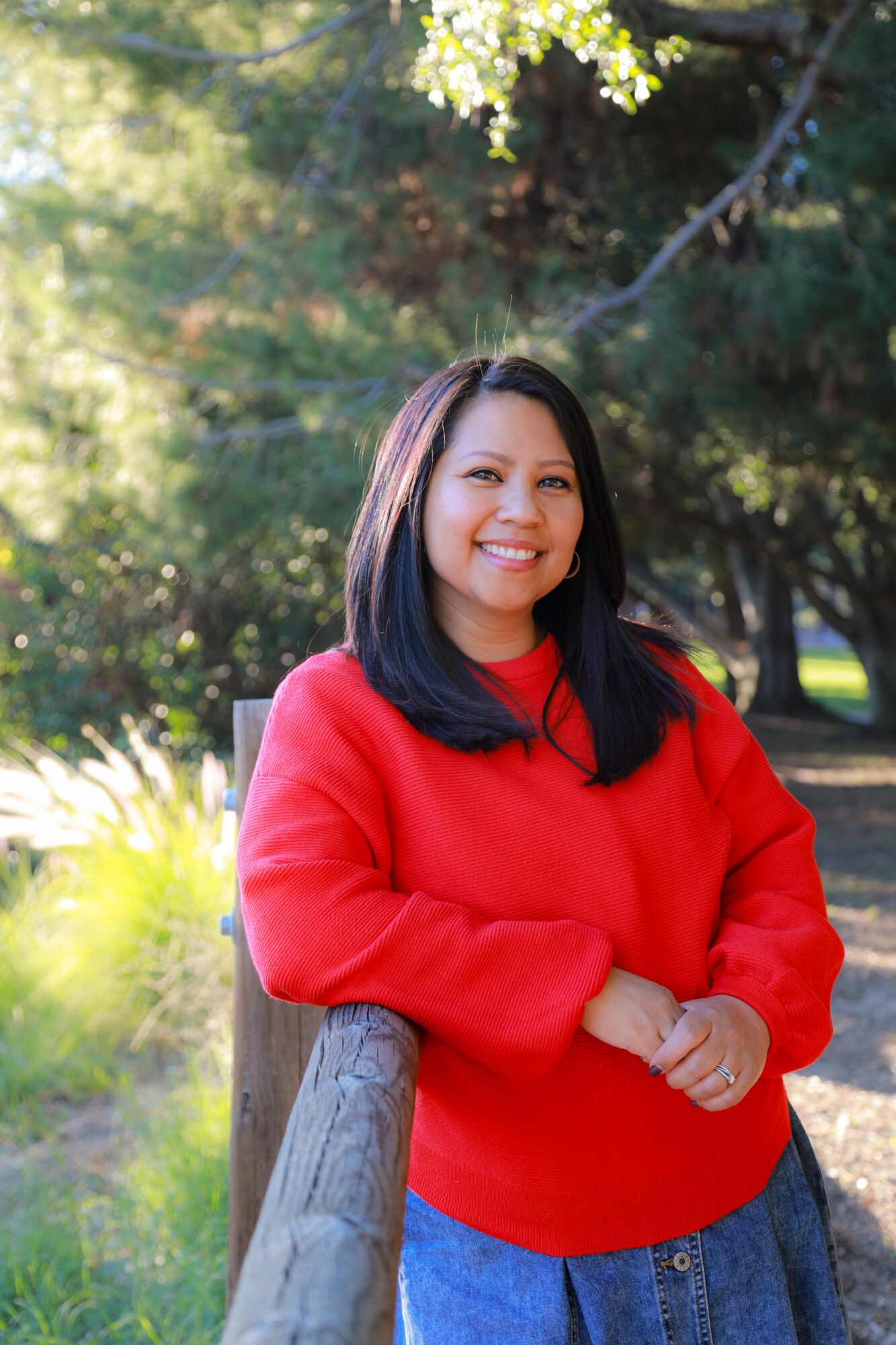 Smiling woman with shoulder-length black hair wearing a red sweater outdoors, leaning on a wooden railing, surrounded by trees and greenery.
