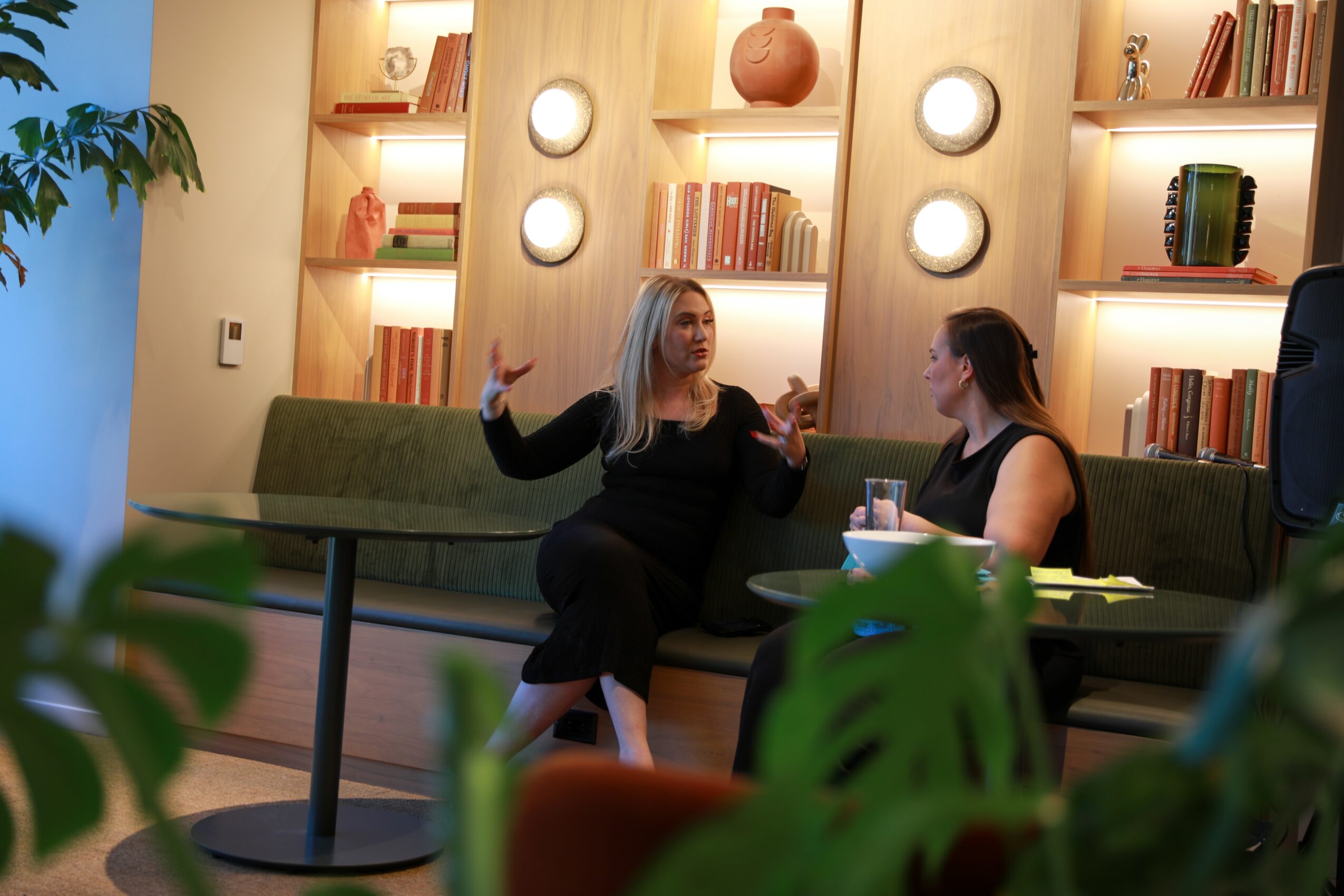 Two women sit on a green sofa in a room with wooden shelves and books, engaged in conversation, with plants in foreground.