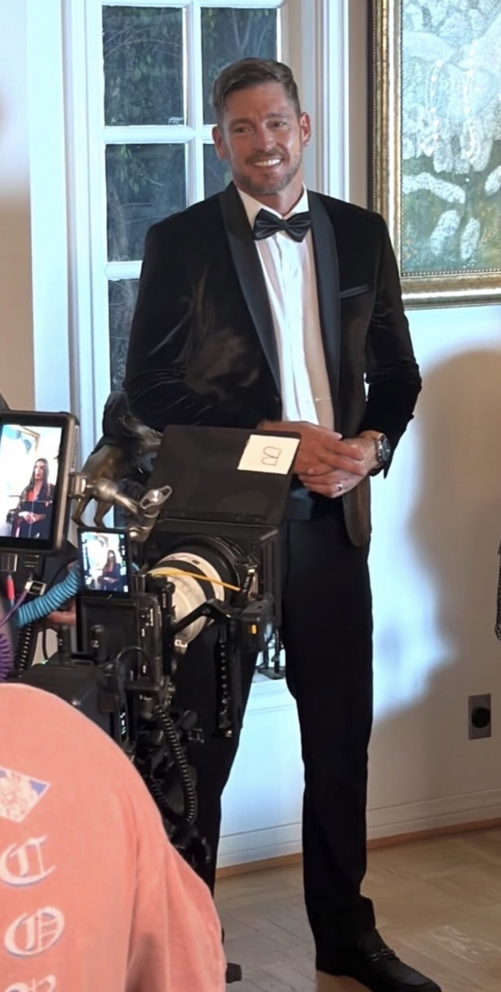 Man in tuxedo holding an award, standing indoors near a window, with camera equipment in foreground.